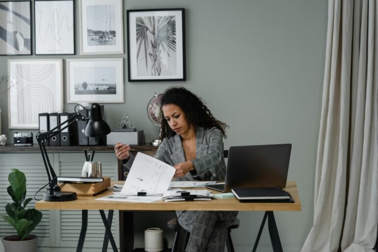 Black woman entrepreneur overwhelmed with paperwork and laptop at desk in home office